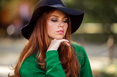 Kırmızı kıvırcık saçlı Sunshine genç genç kız sonbahar parkta yeşil kazak ve şapka giyiyor.