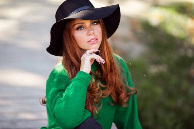 Kırmızı kıvırcık saçlı güneşli genç genç kız Portresi sonbahar parkta yeşil kazak ve moda şapka giyiyor.