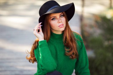 Kırmızı kıvırcık saçlı güneşli genç genç kız Portresi sonbahar parkta yeşil kazak ve moda şapka giyiyor.