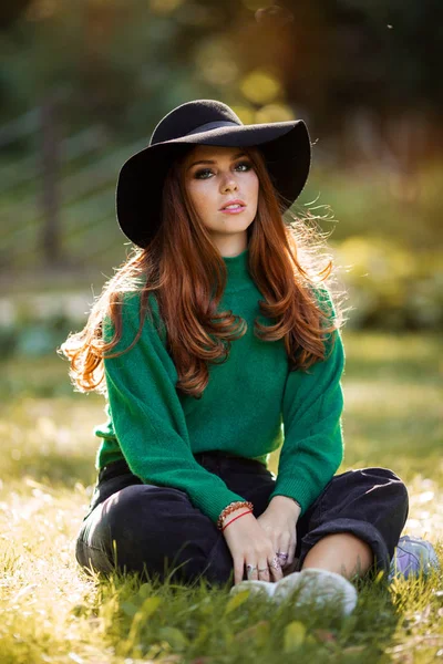 Słońce młody nastolatków dziewczyna z czerwony kręcone włosy ma na sobie zielony sweter z dzianiny i kapelusz siedzi na trawie w jesiennym parku. — Zdjęcie stockowe