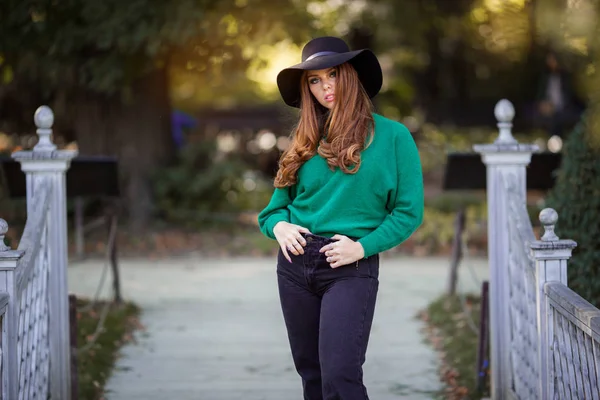 Sluníčko mladá dívka s červenými kudrnatými vlasy nosí zelený pletený svetr a klobouk v podzimním parku. — Stock fotografie