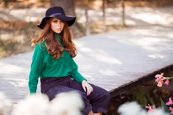 Portrait of sunshine young teen girl with red curly hair is wearing green sweater and fashion hat is sitting on bridge in autumn park. — Stock Photo, Image