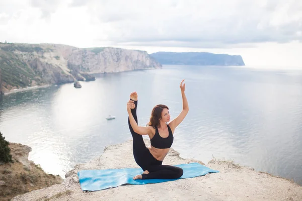 Hermosa mujer delgada practica yoga y pilates en el acantilado de la montaña con vistas al mar. Concepto de estilo de vida deportivo y pagano . — Foto de Stock