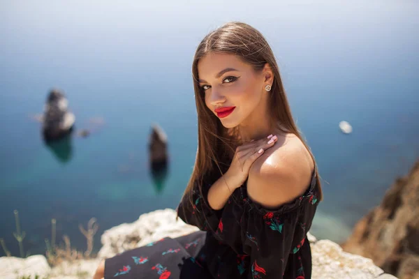 Jovem mulher bonita com maquiagem perfeita está vestindo vestido de moda no penhasco da montanha com a vista azul do mar — Fotografia de Stock