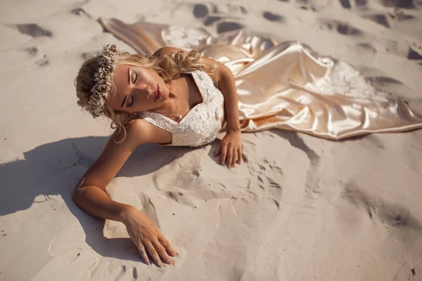 Hermosa novia está usando vestido de vuelo de moda y corona de flores tumbado en la arena en el gran desierto de Dubai . — Foto de Stock