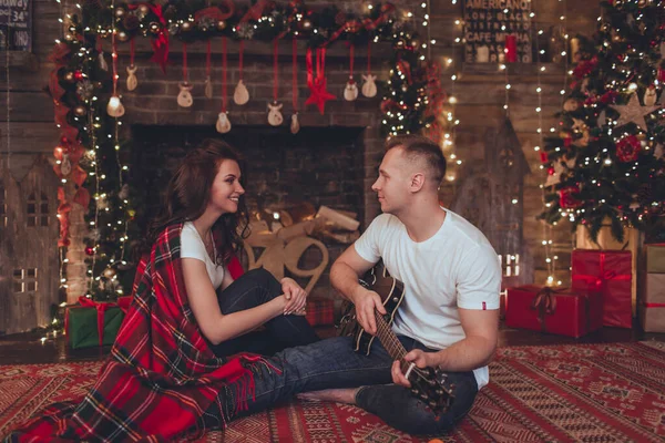 Lovely beautiful couple woman and man are sitting at home near fireplace and playing guitar. Christmas time. — Stok fotoğraf