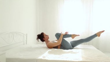 Spor kıyafetli genç bir kadın sabah erkenden beyaz yatak odasında karın kası egzersizi yapıyor..