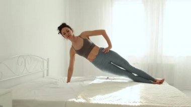 Spor giyimli genç bir kadın, sabahın erken saatlerinde beyaz yatak odasında yoga yapıyor..