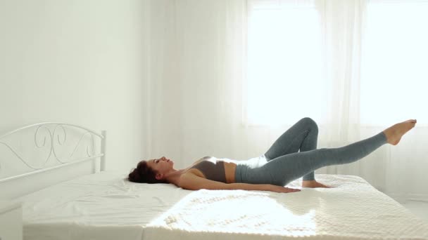 Ung fitness kvinna i sportkläder tränar yoga på sängen i vitt sovrum hemma tidigt på morgonen. — Stockvideo