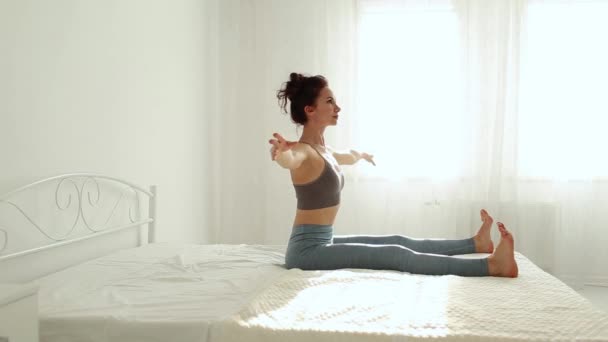Giovane donna fitness in abbigliamento sportivo praticare yoga sul letto in camera da letto bianca a casa al mattino presto . — Video Stock