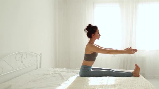 Jeune femme de fitness en vêtements de sport pratiquant le yoga sur le lit dans la chambre blanche à la maison tôt le matin . — Video