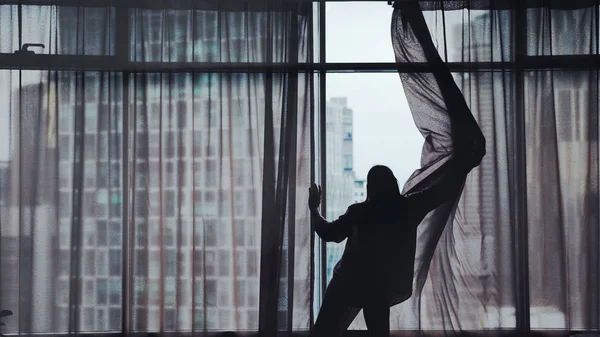 Silhouette of young woman opening window curtains on building on the background