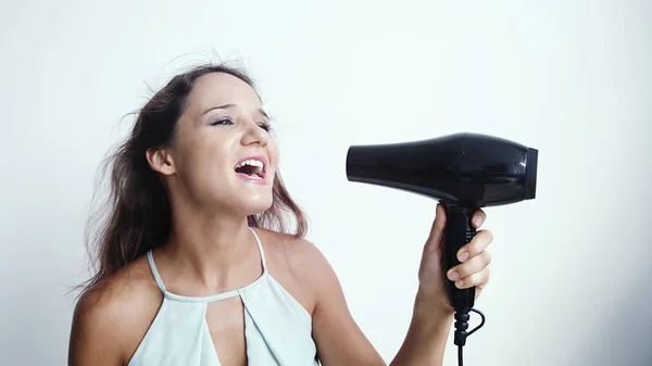 Beautiful young happy woman sings song while using hairdryer on a white background