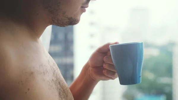 Close up of young handsome man hand with a cup of coffee looks at city towers from a window.