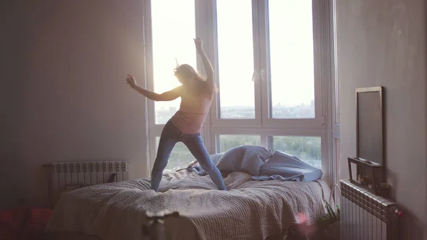 Happy young woman jumps and dances on bed in luxury apartment during beautiful sunrise with lens flare effect in the morning