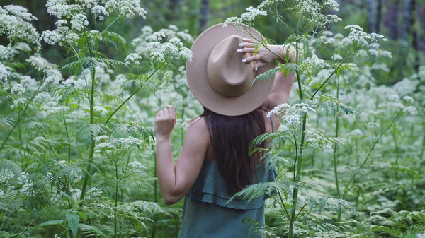 Young pretty woman in hat poses in deep forest among white flowers.