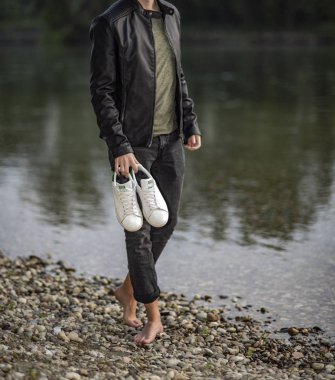 Pavia, Italy - April 30, 2017: Young man barefoot holding a pair of Adidas Stan Smith shoes in his hand near the river - illustrative editorial clipart
