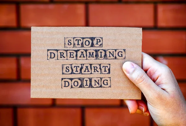 Woman hand holding cardboard card with words Stop Dreaming Start Doing made by black alphabet stamps against brick wall background.