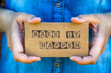Make It Happen siyah alfabe pullar tarafından yapılan kelimelerle karton kartı tutan kadın el. Denim arka planlar. 