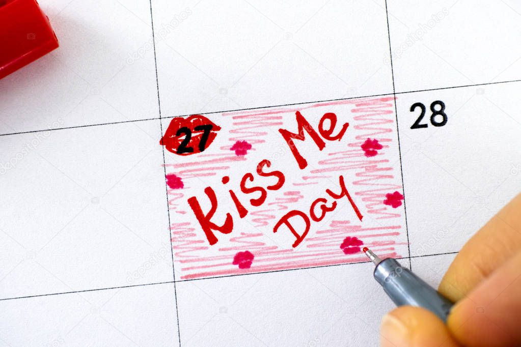 Woman fingers with pen writing reminder Kiss Me Day in calendar.
