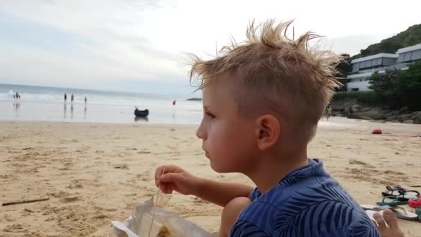 Niño comiendo un panqueque en un fondo de playa con el mar. — Vídeo de stock