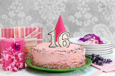Renkli şekerli pembe doğum günü pastası ve 16 numaralı mum. Pasta hediyelerle, tabak ve çiçeklerle çevrili..