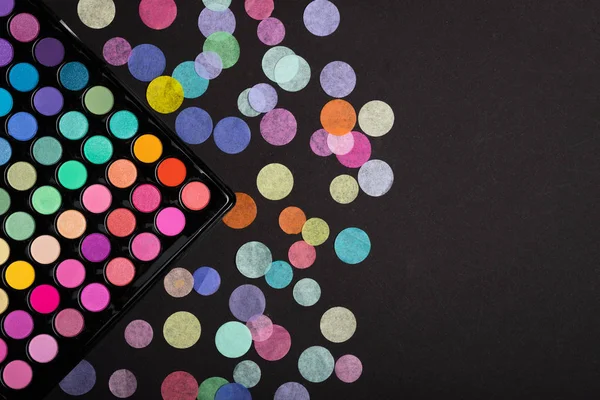 Makeup eyeshadow palette with colorful confetti scattered on black background