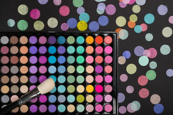 Cepillo de maquillaje en la paleta de maquillaje con confeti de colores sobre fondo negro —  Fotos de Stock