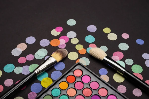 Two brushes with eyeshadow palette on back background decorated with colorful confetti — Stock Photo, Image