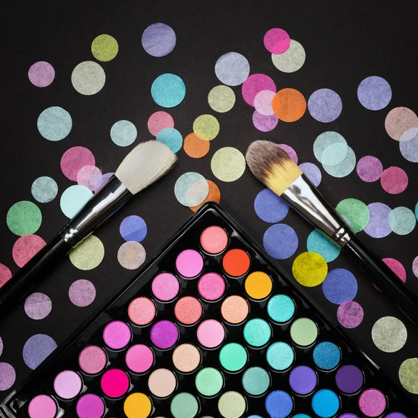 Pinceles cosméticos con paleta de sombras de maquillaje sobre fondo negro con confeti —  Fotos de Stock