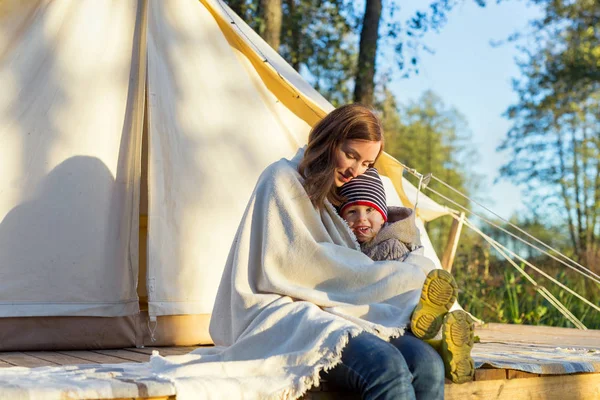 Happy mother embracing her kid with a blanket while sitting near canvas tent — Stock Photo, Image