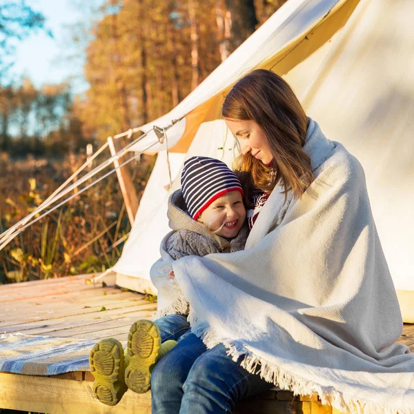 Happy mother embracing her kid with a blanket while sitting near canvas tent — Stock Photo, Image