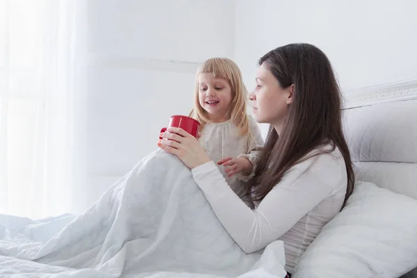 白い寝室でベッドに座りながらお茶を飲む娘を持つ若い母親 — ストック写真