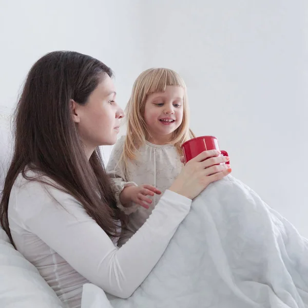 Junge Mutter mit Tochter trinkt Tee, während sie im Bett im Schlafzimmer sitzt — Stockfoto