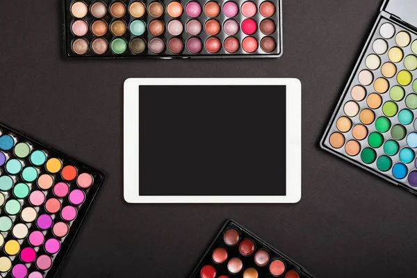 Touchpad with blank screen with colorful eyeshadow palettes on black background. Flat lay