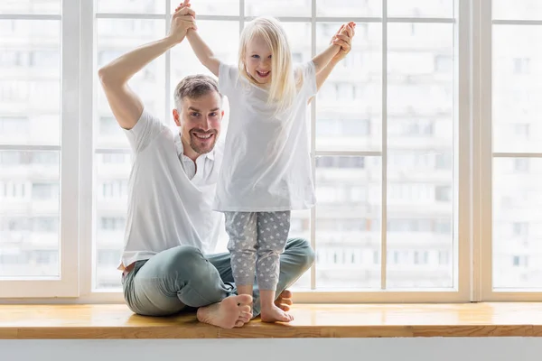 Feliz padre sentado en el alféizar de la ventana con su hija de 3 años con las manos levantadas —  Fotos de Stock