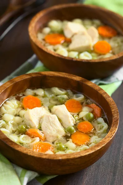 Ev yapımı tavuk çorbası bezelye, havuç ve ahşap kase, küçük kabuk makarna ile fotoğraflandı doğal ışık (seçici odak, odak ilk çorba içinde tavuk önündeki ile)
