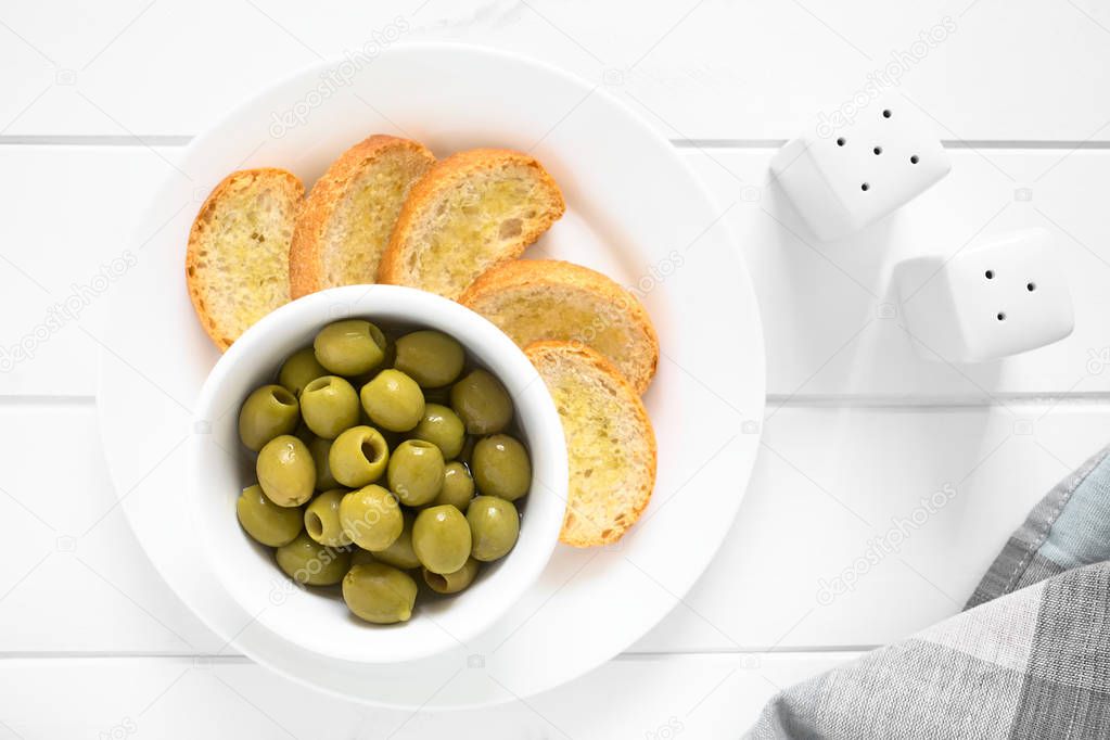 Pitted green olives in bowl with crostini on the side, photographed overhead on white wood with natural light
