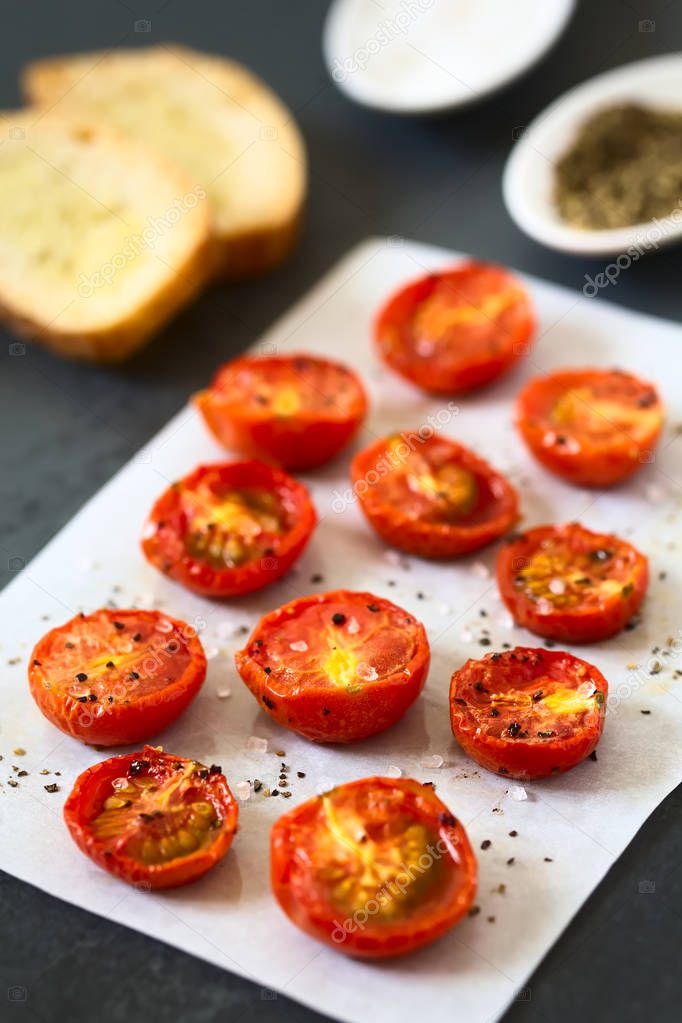 Roasted cherry tomato halves with salt and pepper on baking paper, crostini and spices in the back, photographed with natural light (Selective Focus, Focus one third into the image)