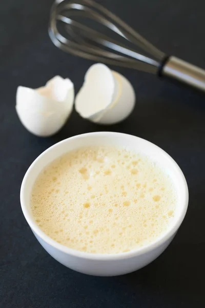 Whisked whole eggs (whites and yolks together) in bowl, with whisk and egg shells in the back, photographed on slate with natural light (Selective Focus, Focus in the middle of the whisked egg)