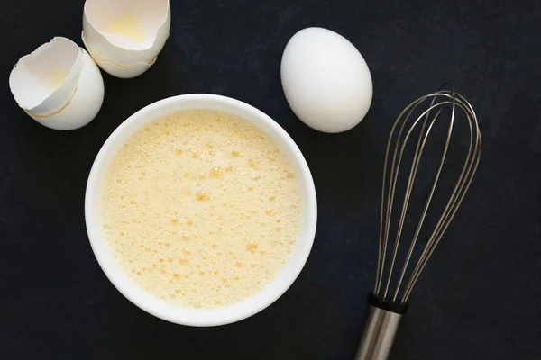 把整个鸡蛋 白色和蛋黄放在一起 放在碗里 旁边有搅拌和蛋壳 在头顶上拍摄自然光 — 图库照片