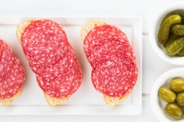 Tarafında yeşil zeytin ve gherkins ile ciabatta ekmek salam dilimleri, beyaz ahşap üzerinde havai fotoğraflandı (Seçici Odak, Salam dilimleri odaklanın)