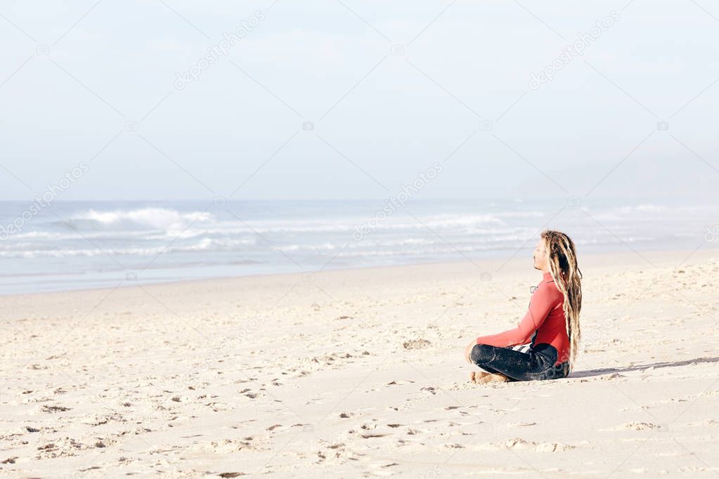 Young adult surfer with dreadlocks wearing wetsuit sitting cross-legged on beach near spot and meditating before summer evening surfing session - yoga and surfing concept. Baleal, Peniche, Portugal
