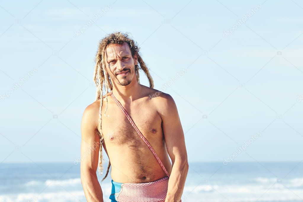 Portrait of young adult shirtless surfer with dreadlocks standing and smiling on beach against surfing spot before summer evening session - surfing and extreme sport concept. Baleal, Peniche, Portugal