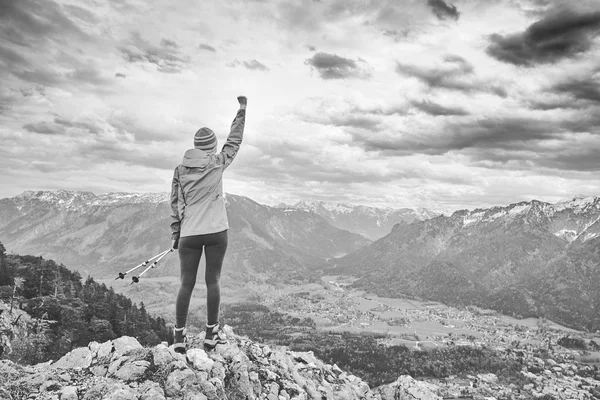 Black and white portrait of young female hiker with trekking poles and arm raised celebrating achievement standing on top of mountain after successful ascent - adventure, freedom or success concept