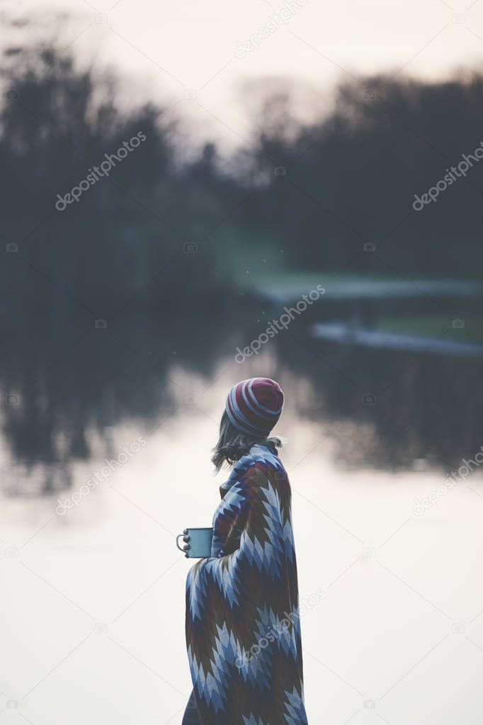 Vintage color portrait of young blonde woman in red cap and knitted colourful blanket enjoying sunset silence near lake with mug of tea - peace, calm and serenity concept