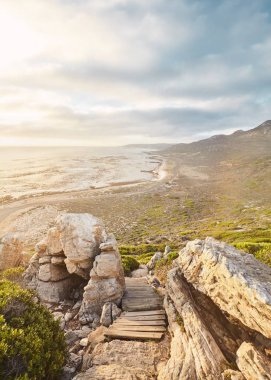 Stunning view through boulders and wooden footpath to ocean and mountains on golden sunset near Cape of Good Hope, South Africa
