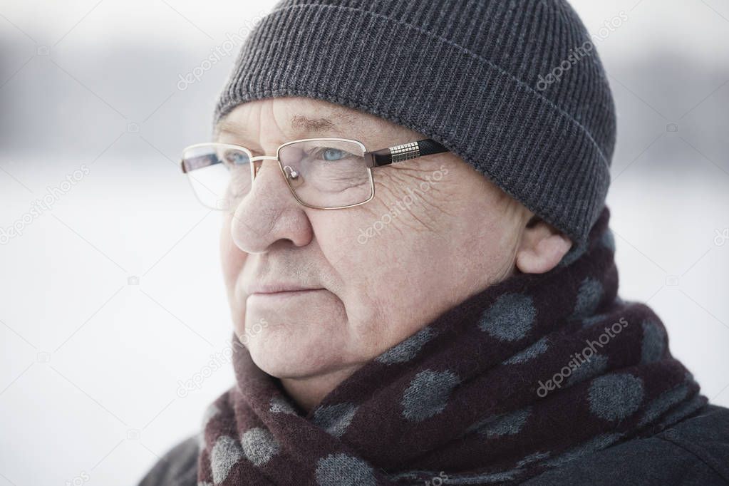 Close up portrait of senior man wearing glasses, knit cap and scarf standing outdoors in winter