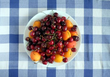Sağlıklı beslenme. Damalı masa örtüsü üst görünümü closeup olgun kayısı ve kiraz dolu beyaz yuvarlak plaka ile natürmort