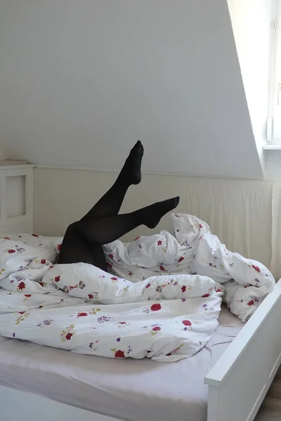 Sexy female legs in black pantyhoses look above up from under the blanket in uncovered white bed against the wall